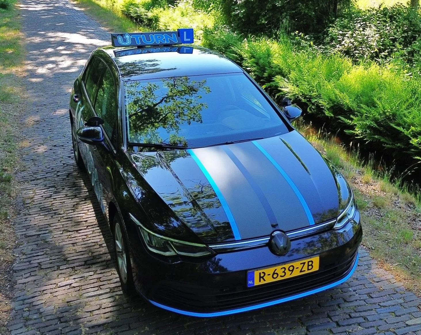 Zwarte rijschoolauto met blauwe strepen in bosrijke omgeving van voren